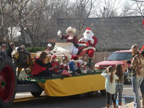 A festive parade float featuring Santa and a woman in a holiday outfit, surrounded by children and decorations.