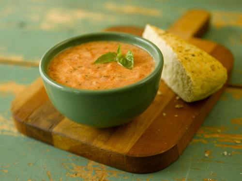 A bowl of creamy tomato soup garnished with basil, served with a piece of bread on a wooden cutting board.