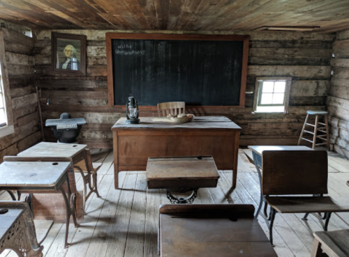 A rustic classroom with wooden desks, a chalkboard, and a teacher's desk, featuring a lantern and a portrait on the wall.
