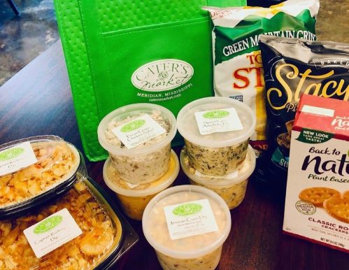 A variety of packaged foods, including dips, snacks, and a green tote bag from a local catering service.