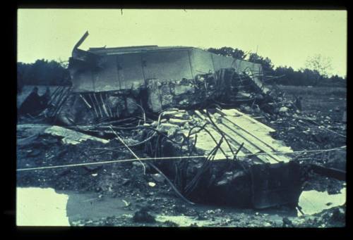 A collapsed structure surrounded by debris and mud, with trees in the background under a cloudy sky.