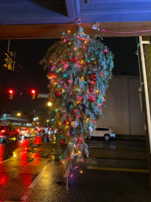 A large, upside-down Christmas tree adorned with colorful lights and ornaments, hanging in a rainy urban setting.