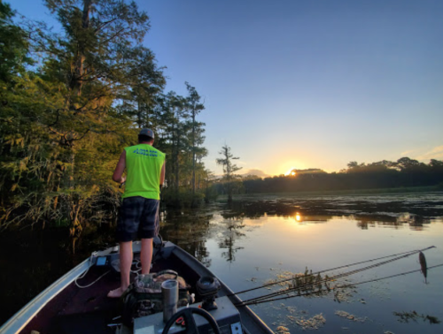 A person fishing from a boat at sunset, surrounded by trees and calm water reflecting the colorful sky.