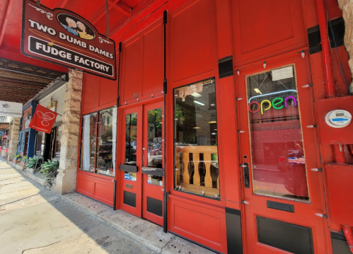 Exterior of a red storefront with a sign reading "Two Dumb Dames Fudge Factory" and an "Open" neon sign.