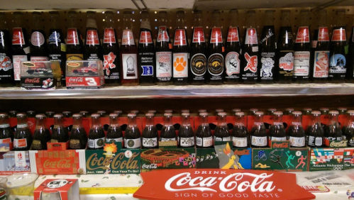 A display of vintage Coca-Cola bottles and memorabilia on a shelf, showcasing various designs and packaging.
