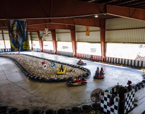 Indoor go-kart track with racers navigating curves, surrounded by tires and colorful banners.