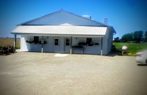 A white metal building with a sloped roof, surrounded by gravel and green fields under a clear blue sky.
