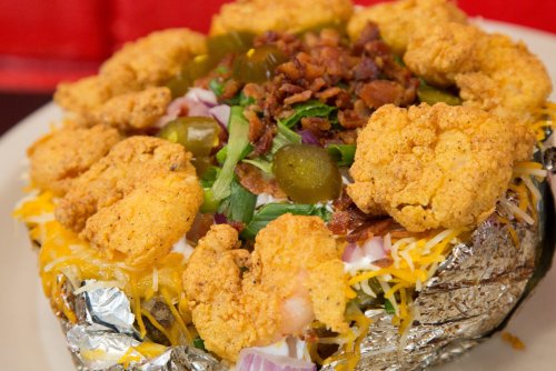 A loaded baked potato topped with fried chicken, cheese, bacon, jalapeños, and green onions, wrapped in foil.