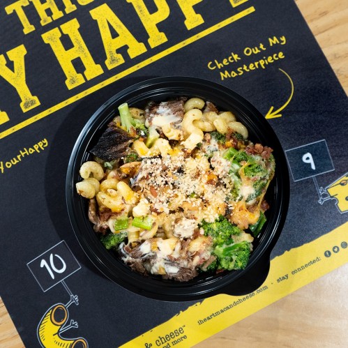 A black bowl filled with macaroni, cheese, broccoli, and toppings, placed on a colorful menu background.
