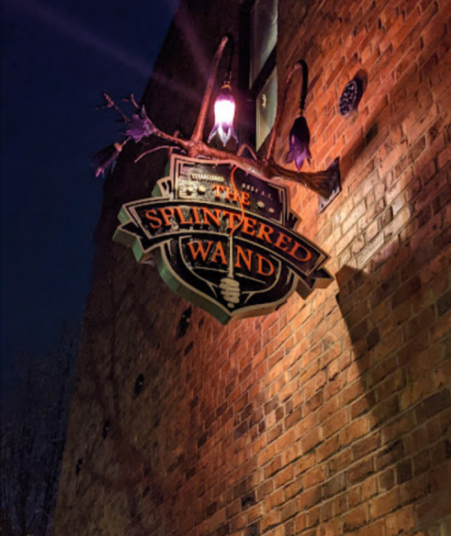 Sign for "The Splintered Wand" hanging on a brick wall, illuminated by a lantern at night.