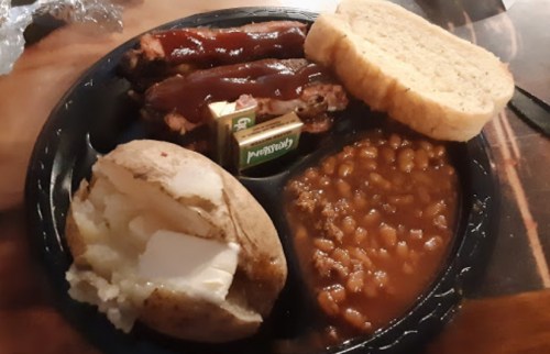 A plate with barbecue ribs, baked beans, a baked potato with butter, and slices of bread.
