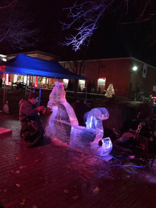 A sculptor carves an ice sculpture of a horse and sleigh at night, illuminated by colorful lights.