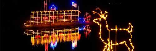 Colorful holiday lights illuminate a boat and a reindeer silhouette, reflecting on the water at night.