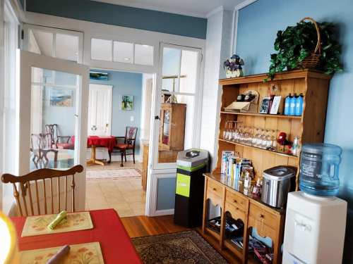 A cozy dining area with a wooden table, shelves of glassware, and a water cooler, leading to a bright room.