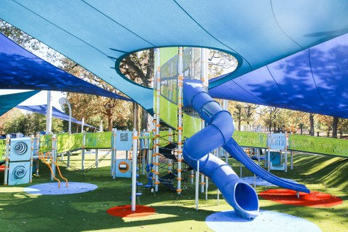 Colorful playground with a blue slide, climbing structures, and shaded areas under a bright blue sky.