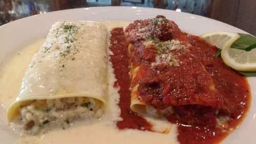 Two enchiladas on a plate: one with creamy white sauce and the other with red sauce, garnished with herbs and a lemon wedge.