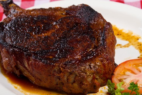 A perfectly cooked, charred ribeye steak on a plate, garnished with a slice of tomato and herbs.