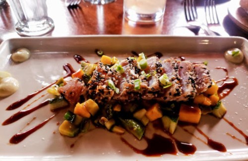 A plate of sesame-crusted fish on a bed of diced mango and cucumber, drizzled with sauce and garnished with green onions.