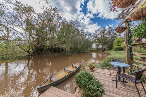 A serene riverside scene with a canoe, wooden deck, and chairs, surrounded by trees and a cloudy sky.