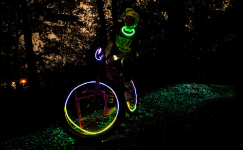 A cyclist in neon gear rides a bike with glowing wheels on a dark, tree-lined path.