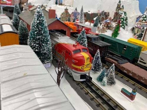 A model train set featuring a red locomotive, surrounded by miniature trees and buildings in a snowy landscape.