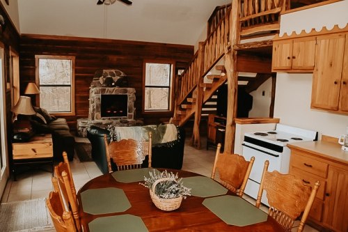 Cozy cabin interior with wooden furniture, a stone fireplace, and a dining table in a warm, inviting atmosphere.