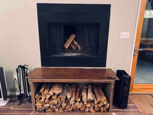 A fireplace with logs stacked below, set against a light-colored wall, with a wooden table and a black heater nearby.