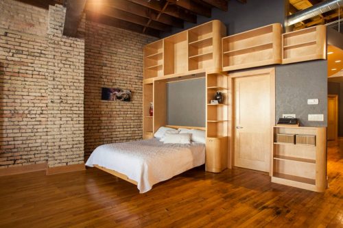 A modern bedroom with a wooden bed frame, exposed brick walls, and built-in shelves, featuring warm wooden flooring.