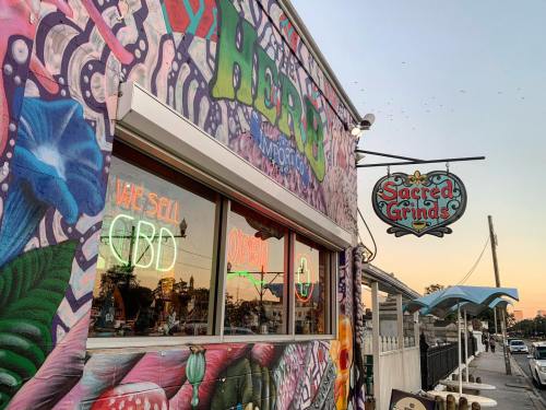Colorful mural on a building with a sign for "Sacred Grinds" and neon lights, set against a sunset sky.
