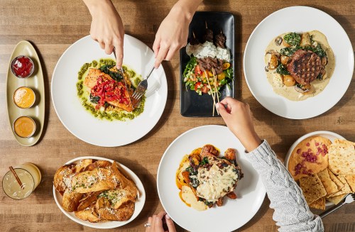 A table spread with various dishes, including pasta, steak, and salads, with hands reaching for the food.