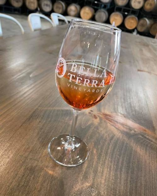 A glass of rosé wine with the logo of Bella Terra Vineyards, set on a wooden table with barrels in the background.