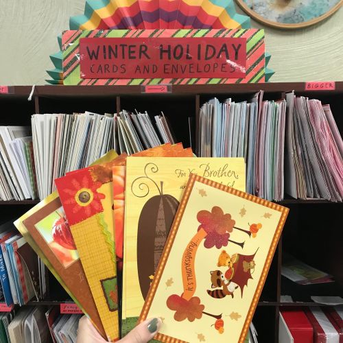A hand holds colorful winter holiday cards in front of a shelf filled with various envelopes and cards.