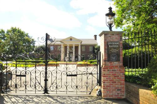A grand entrance gate with ornate ironwork leads to a stately mansion surrounded by lush greenery.
