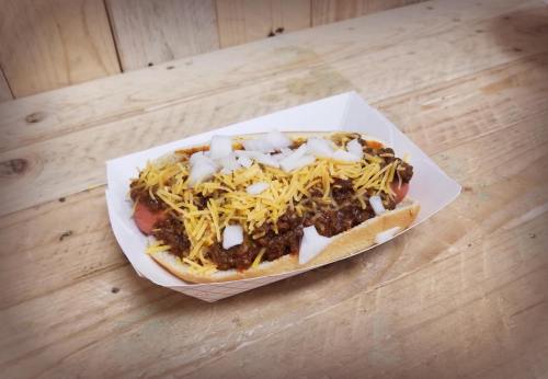 A hot dog topped with chili, shredded cheese, and diced onions, served in a paper tray on a wooden surface.