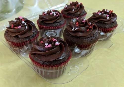 A clear container holding six chocolate cupcakes with swirled frosting and heart-shaped sprinkles on top.