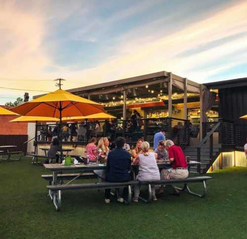 Outdoor dining area with people at picnic tables, yellow umbrellas, and a building under a colorful sunset sky.