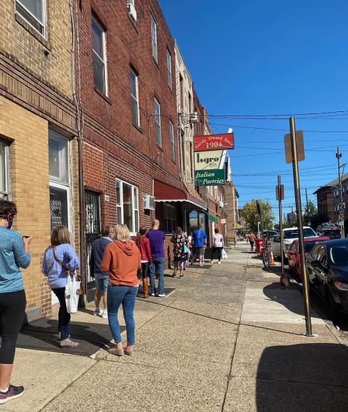 A long line of people waiting outside an Italian market on a sunny day, with brick buildings and parked cars nearby.