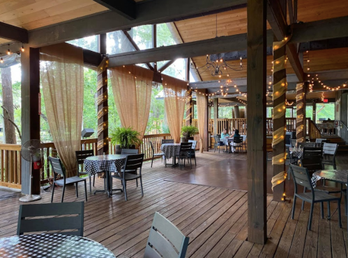 A cozy restaurant patio with wooden floors, checkered tables, and string lights, surrounded by greenery.