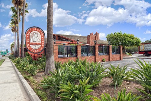 Exterior view of a restaurant with a brick facade, palm trees, and a sign, set against a blue sky with clouds.