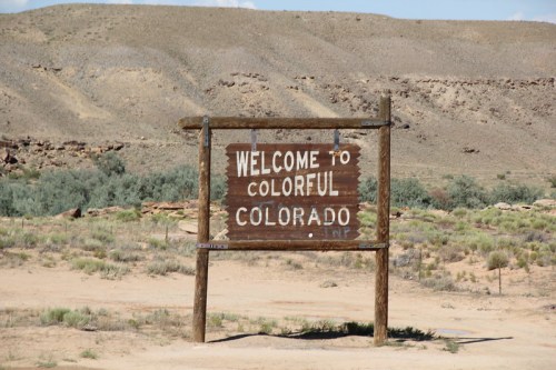 A wooden sign reading "Welcome to Colorful Colorado" in a desert landscape with sparse vegetation and hills in the background.