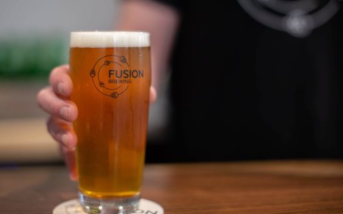 A person holding a pint of beer with a "Fusion Brewing" logo on the glass, set on a wooden bar.
