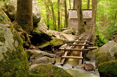 A wooden cabin nestled among trees and rocks, connected by a bridge over a flowing stream in a lush forest.