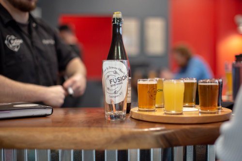 A wooden bar with a bottle of Fusion Brewing and a flight of various beer samples in small glasses.