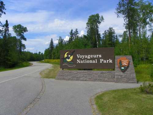 Sign for Voyageurs National Park at the entrance, surrounded by trees and a clear blue sky. Road leads into the park.