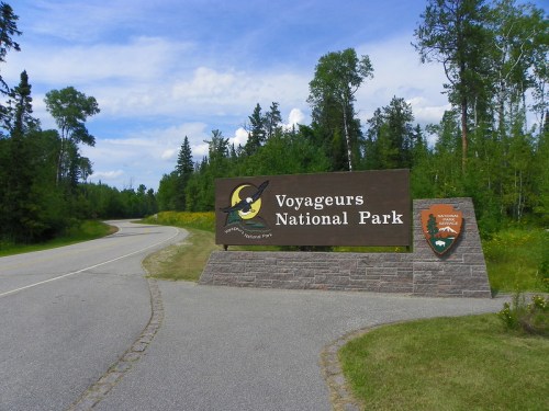 Sign for Voyageurs National Park at the entrance, surrounded by trees and a clear blue sky. Road leads into the park.