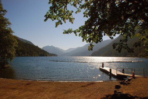 A serene lake surrounded by mountains, with a wooden dock extending into the water under a clear blue sky.