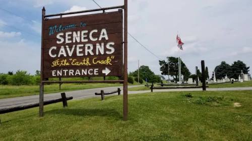 Sign for Seneca Caverns with "Entrance" arrow, surrounded by green grass and trees under a cloudy sky.