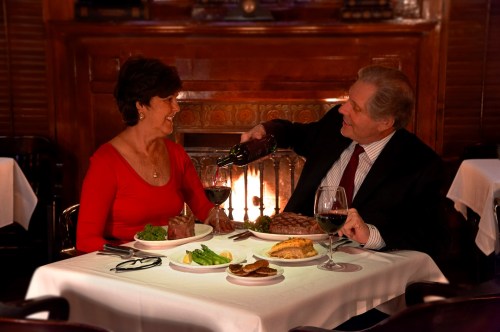 A couple enjoys a romantic dinner by a fireplace, sharing wine and a meal at an elegant restaurant.