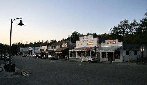 A quiet street lined with small shops and buildings at dusk, with trees in the background and a few parked cars.