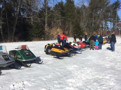 A group of people gathered around parked snowmobiles on a snowy landscape, surrounded by trees.